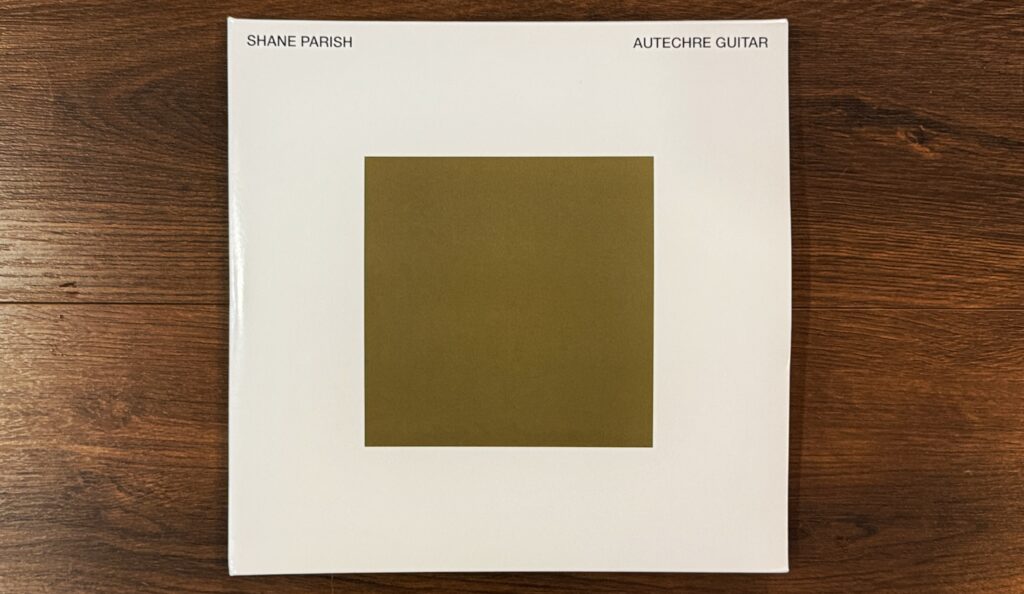 LP copy of the album on a wood floor. The art is a solid white background with a smaller solid gold/brown square centered in the middle with smaller text SHANE PARISH in the upper left corner and AUTECHRE GUITAR in the upper right.