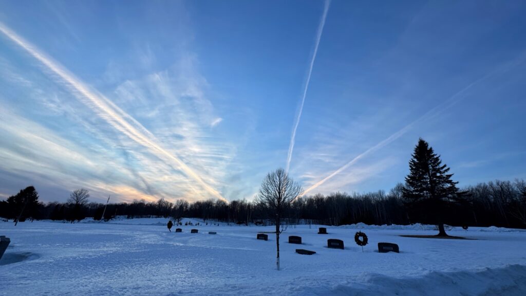 Several white lines in the sky seem to converge on one point over the horizon in an otherwise clear blue sky. The ground is covered in crusty snow.